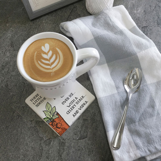 Bloody Mary coaster on a gray stone surface beside a latte cup with latte art, spoon and striped napkin.