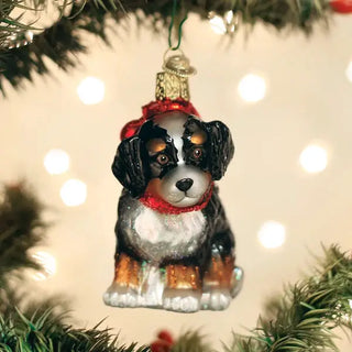 Bernedoodle puppy ornament, black and tan glass figure with white chest and red scarf, hanging on a Christmas tree.