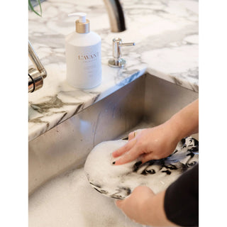 Fresh Linen Dish Soap by L’AVANT Collective on marble counter, hand washing a foamy plate.