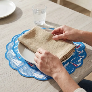 Placemat set of 4 nautical blue scalloped placemats with sailboats and lighthouses, beige napkin on white plate.