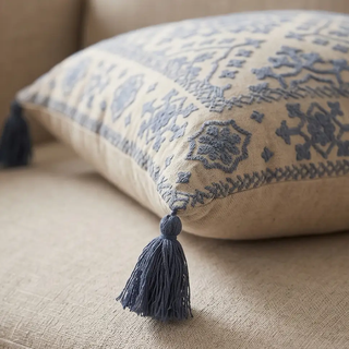Square pillow handcrafted in beige with blue embroidery and tassels on corners, featuring a geometric motif
