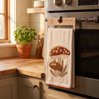 Mushroom dish towel hanging on the oven door, cream fabric with an orange-brown mushroom print.
