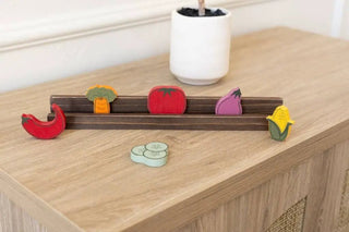 Six wooden veggie shapes on a brown display rack: carrot, tomato, corn, eggplant, red pepper, all on a light wood table.