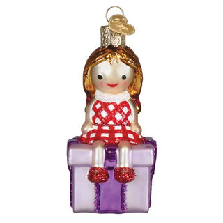 Misfit Doll Ornament; girl figure in red plaid dress sits on a purple gift box with a gold hat accent
