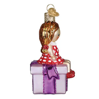 Misfit Doll Ornament in red-and-white check dress with gold hair, perched on a purple gift box.