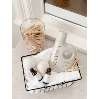 Laundry detergent bottle in soft cream with gold lettering, Fresh Linen scent, in a basket with two bottles and towels.
