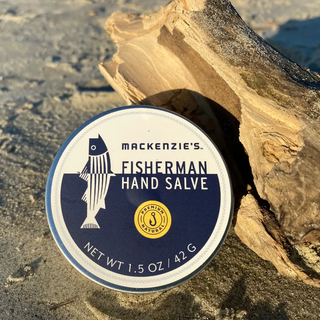 Fisherman Hand Salve tin by Mackenzie’s with blue-and-white label on a sandy beach with driftwood