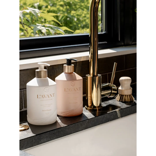 Two dish soap bottles in white and blush pink with pump tops on countertops beside a gold faucet, Blushed Bergamot.