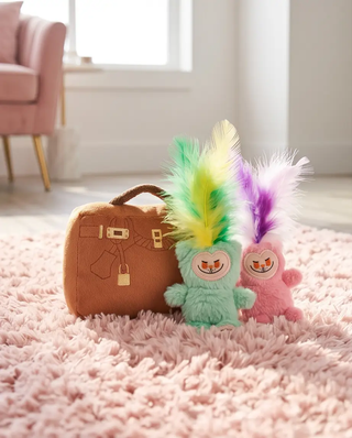 Lameowow handbag catnip cat toy with feathers, two pastel mint and pink plush cats beside a brown bag on a pink shag rug