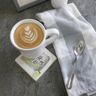 Coaster white ceramic with gin quote under a white mug of latte on a gray countertop; spoon nearby.