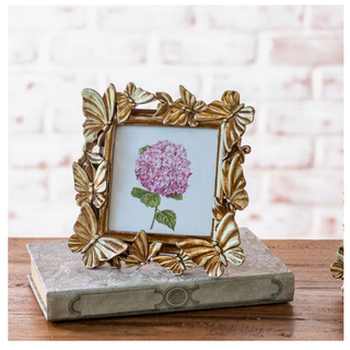 Bronze butterfly frame with leaf motifs housing a pink hydrangea illustration on a decorative gray box.