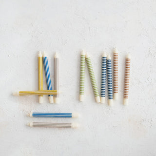 Assorted striped taper candles in blue, yellow, green, gray, and brown, laid out on a light textured surface.