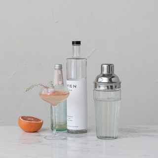 Glass cocktail shaker with stainless steel top on a marble bar, beside a pink cocktail in a coupe, grapefruit half, and two bottles.