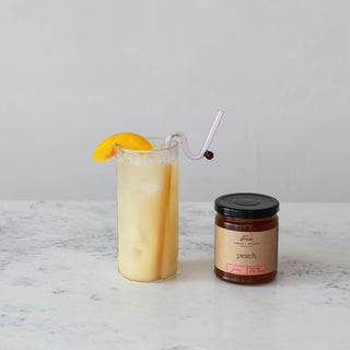 9 oz Peach Jam jar beside a peach cocktail in a tall glass with ice and a peach wedge garnish