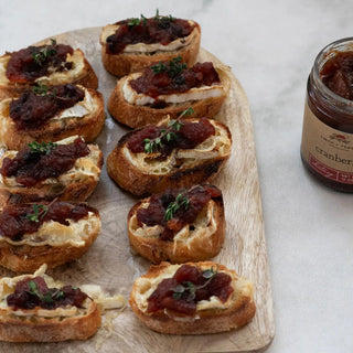 Cranberry chutney crostini bites on toasted baguette rounds with thyme, arranged on a wooden board beside a chutney jar.