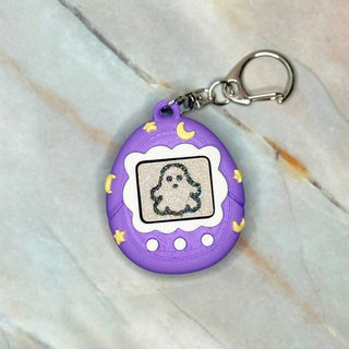 Purple Tamagotchi-style keychain with ghost design on a marble surface