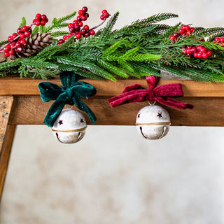 Two white and gold jingle bells with star cutouts, hanging from evergreen garland with red berries.