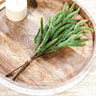Evergreen pick with pine sprigs tied in twine on a round wooden tray beside a white candle.