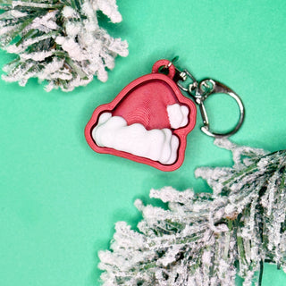 Red Santa hat keychain with white pom-pom on a green background with snow-covered branches.