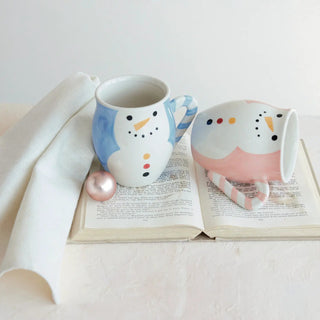 Snowman hand-painted stoneware mugs (2) with blue striped handle and pink candy-cane handle, displayed on an open book.