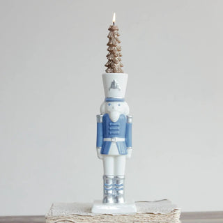 Ceramic nutcracker taper candle holder in white and blue with silver accents, topped by a gold pine-tree candle.
