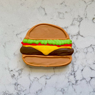 Hamburger-shaped dog biscuit with brown buns, green lettuce, red tomato and yellow cheese.