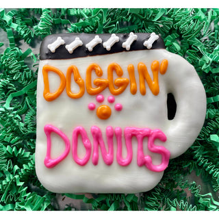 DOGGIN’ DONUTS coffee dog treat cookie with white icing, orange DOGGIN’, pink DONUTS lettering and bone-topped trim