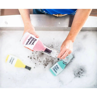 Scrubbly kitchen sponge Rosé Rinse Repeat, pink sponge on a glass bottle; yellow and teal cleaner bottles visible.