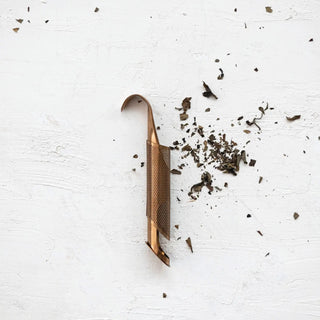 Stainless steel loose tea infuser with over-mug hook in rose-gold finish, perforated body with scattered tea leaves.