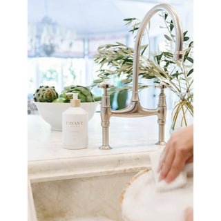 Dish soap bottle with pump on marble kitchen counter; white container with gold-accent label beside a stainless faucet.