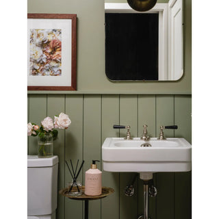 Reed diffuser Blushed Bergamot bottle with pink glass and black reeds on a round wooden table beside a bathroom sink