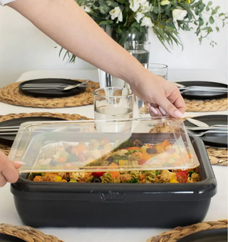 Serving tray with clear lid being lifted to reveal colorful pasta salad.